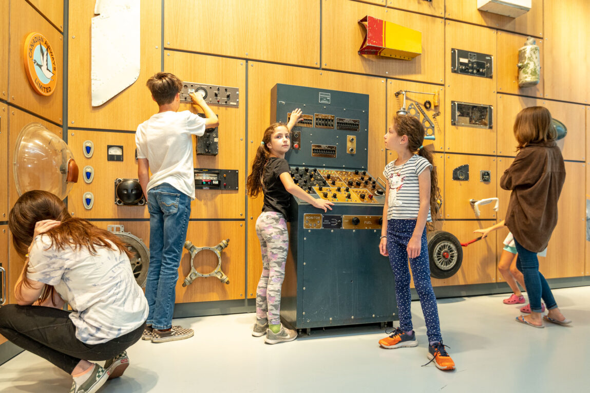 Three children explore the Mechanics' Workshop gallery at the Royal Aviation Museum, playing with radios, dials, and engine simulators.