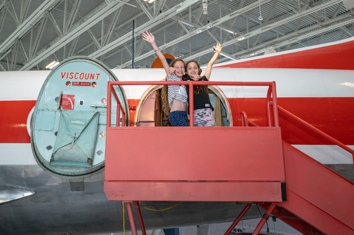 two young girls hold their arms up in excitement as they stand the stairs that lead into the door of a vintage airplane inside a museum