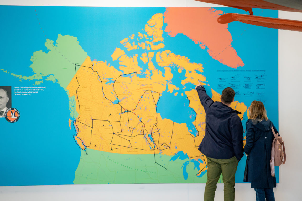 two museum-goers stand in front of a large map of Canada; one is pointing to an area in the Arctic
