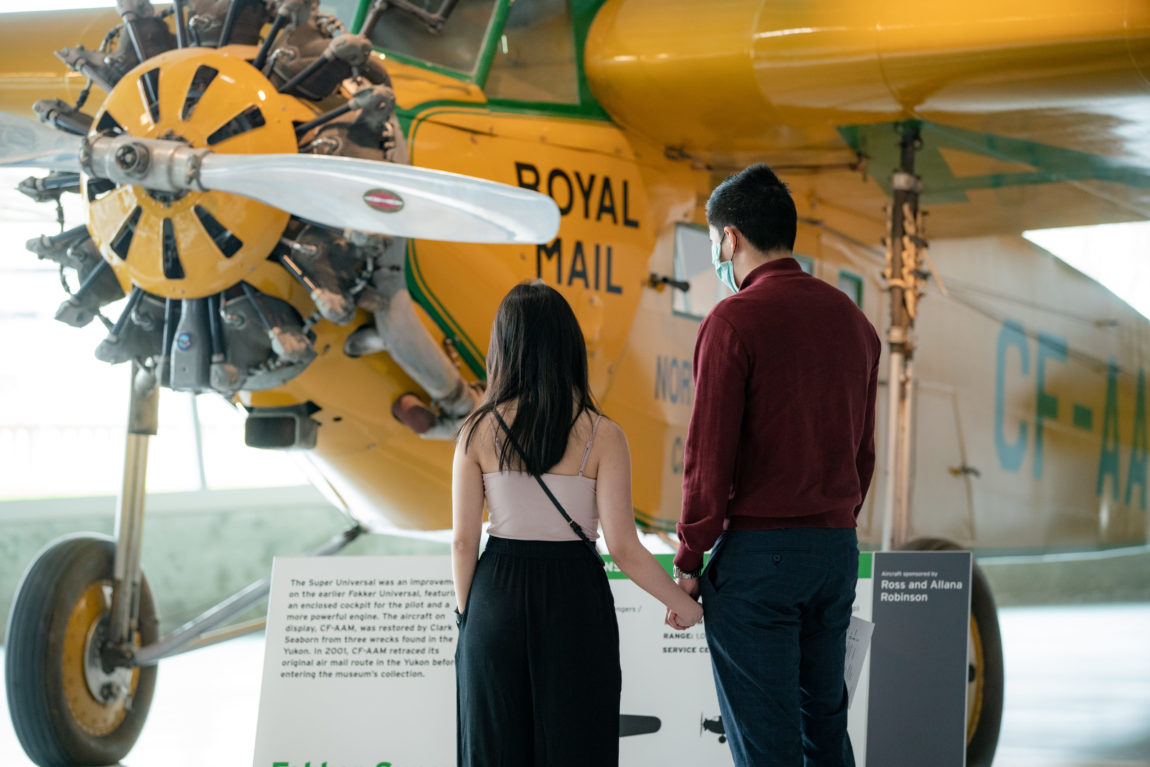 a young couple holds hands while reading the exhibit panel for an aircraft inside an aviation museum