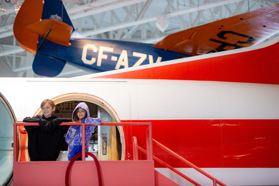 two young children stand on the stairs that lead into the door of a vintage airplane inside a museum