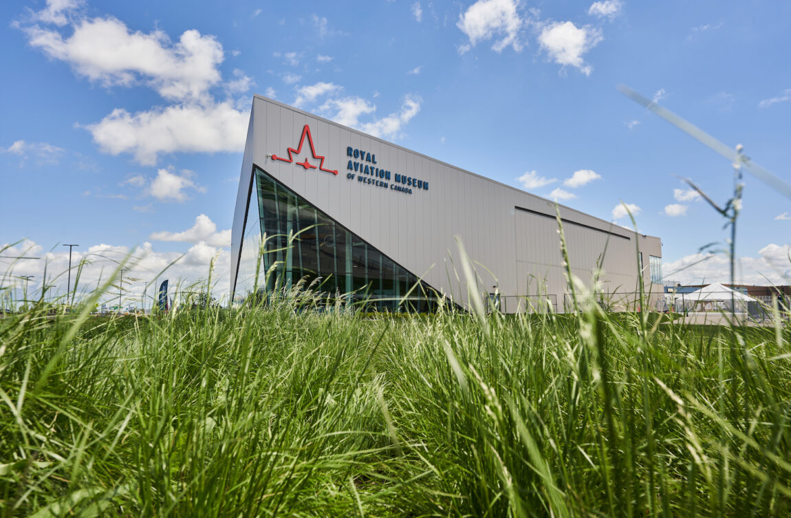 The Royal Aviation Museum of Western Canada, a modern beige building with a glass front peak is pictured against a blue sky dotted with clouds. The museum is photographed from a low angle so the grass in the foreground fills the lower half of the frame.