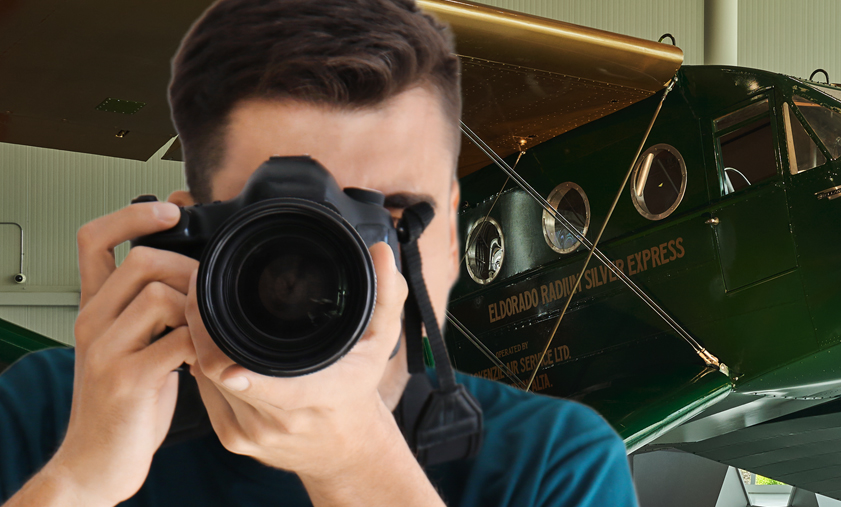 Close-up of someone holding a camera to their eye and pointing it at the camera taking their photo. In the background, the Bellanca Aircruiser, CF-AWR