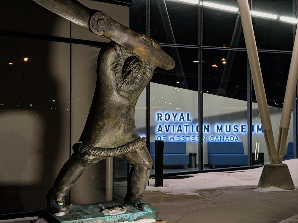 Tom Lamb statue outside the main entrance to the Royal Aviation Museum. Statue is positioned in the first third of the image. To its right, the Royal Aviation Museum logo is visible through windows on a white wall in the museum's lobby.