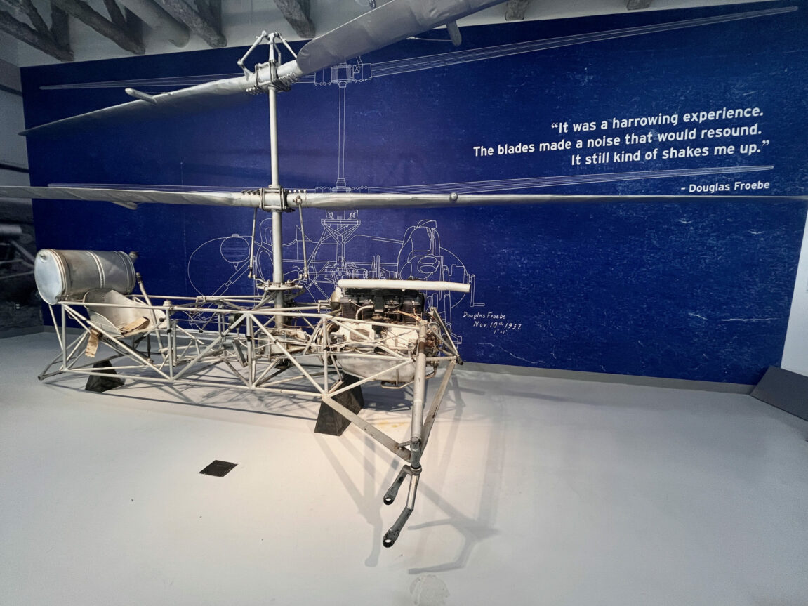 Froebe helicopter on display at the Royal Aviation Museum. The wall behind it is blue and on the wall is a quote from Douglas Froebe that reads, "It was a harrowing experience. The blades made a noise that would resound. It still kind of shakes me up."