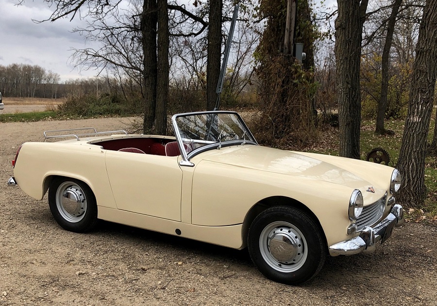 Ivory coloured 1961 Austin-Healey Sprite parked on a gravel road set amidst a forest.