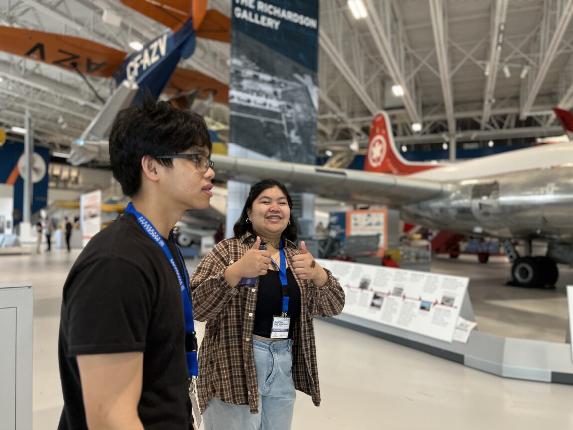 Two high school students stand in the galleries of the Royal Aviation Museum. One faces the camera giving double thumbs-up, the other is in profile, speaking with another student off-camera.