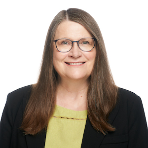 Headshot of a woman with long, straight grey-brown hair, round glasses, wearing a lime green shirt and black blazers set against a white backdrop