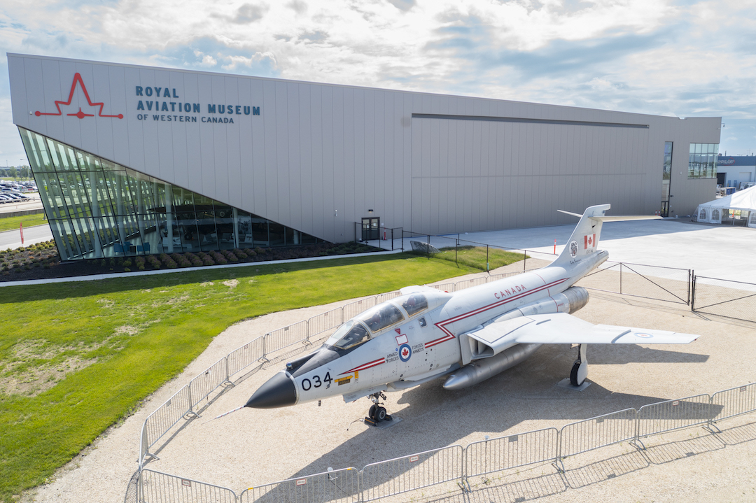 A CF-101 Voodoo fighter jet sits outside a large grey building on the side of which a sign reads, "Royal Aviation Museum of Western Canada"