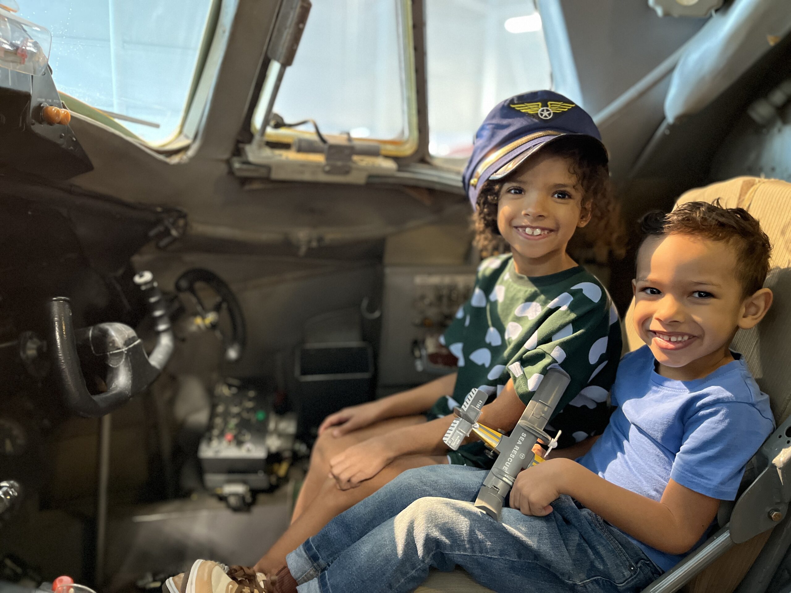 Two young visitors sit in the cockpit of the Royal Aviation Museum's Vickers Viscount. One is wearing an oversized captain's hat. Both are looking at the camera and smiling.