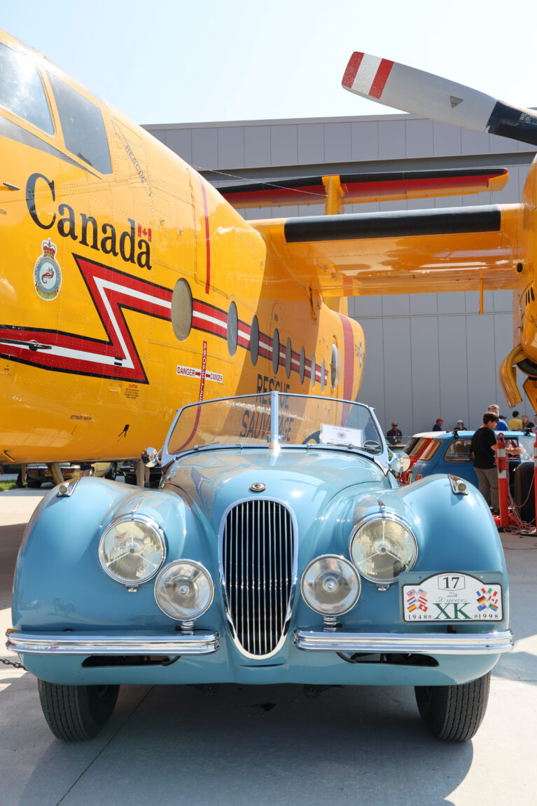 A blue, 1958 Jaguar convertible sits parked next to a CC-115 Buffalo outside of the Royal Aviation Museum of Western Canada