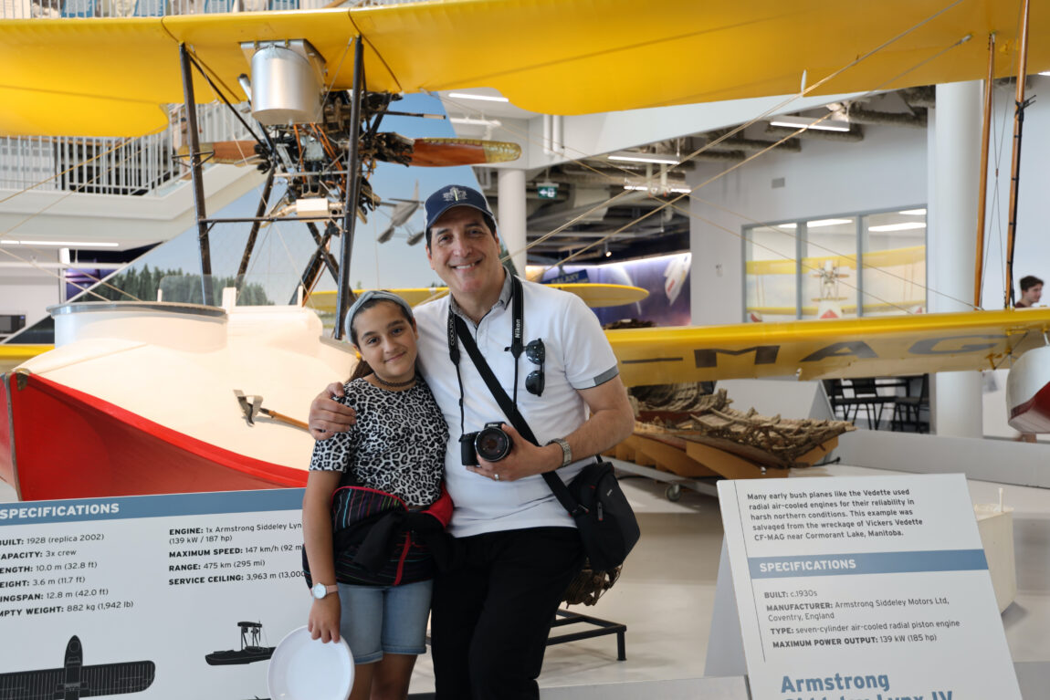 A man wearing a white shirt, black baseball hat, with a camera around his neck poses with his arm around his adolescent daughter who's wearing a floral dress. They are standing in front of a Vickers Vedette aircraft at the Royal Aviation Museum.