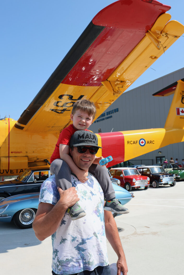 A father wearing sunglasses and a baseball hat smiles at the camera while his young son sits on his shoulders. In the background, part of the Royal Aviation Museum's CC-115 Buffalo is visible.