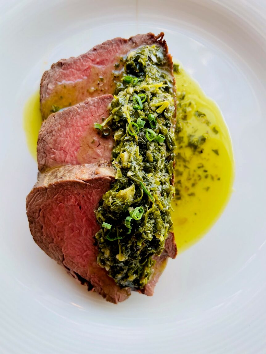 Three pieces of beef tenderloin topped with chimichurri sit layered on a white dinner plate