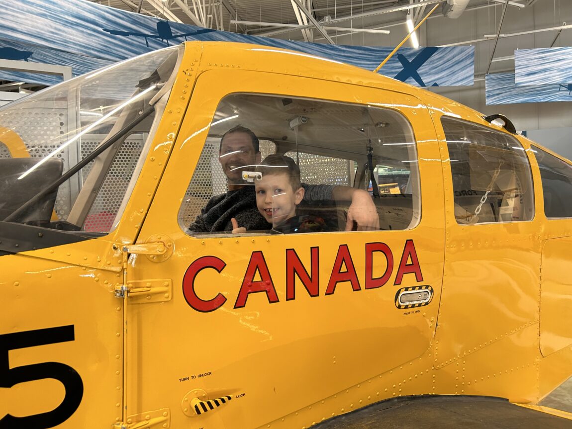 Two visitors sit in the Beechcraft Musketeer at the Royal Aviation Museum. Close-up of the pilot's seat from outside the aircraft. A young boy is looking out the window giving a thumbs up. Visible next to him is a parent figure smiling at the camera.