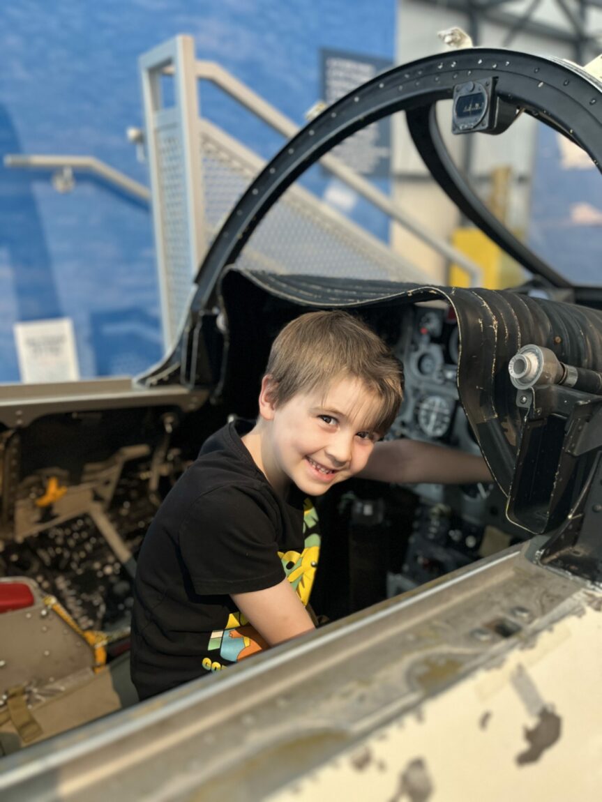 Closeup of a young smiling boy playing in the CF-101 Voodoo flight and tactics trainer at the Royal Aviation Museum. The boy is looking at the camera and his left hand is resting on one of the controls in the cockpit.