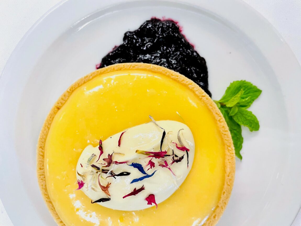A bright lemon tart atop a blackberry coulis sits on a white dinner plate. Photo taken from above.