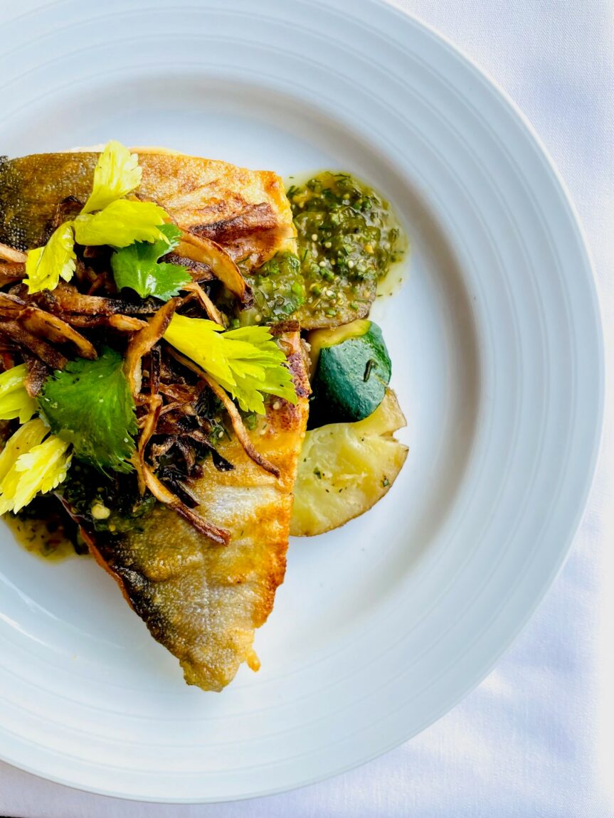 A dinner entree of northern pike garnished with parsley and pesto sits atop a bed of grilled zucchini. The entree is on a white dinner plate, shot from above.