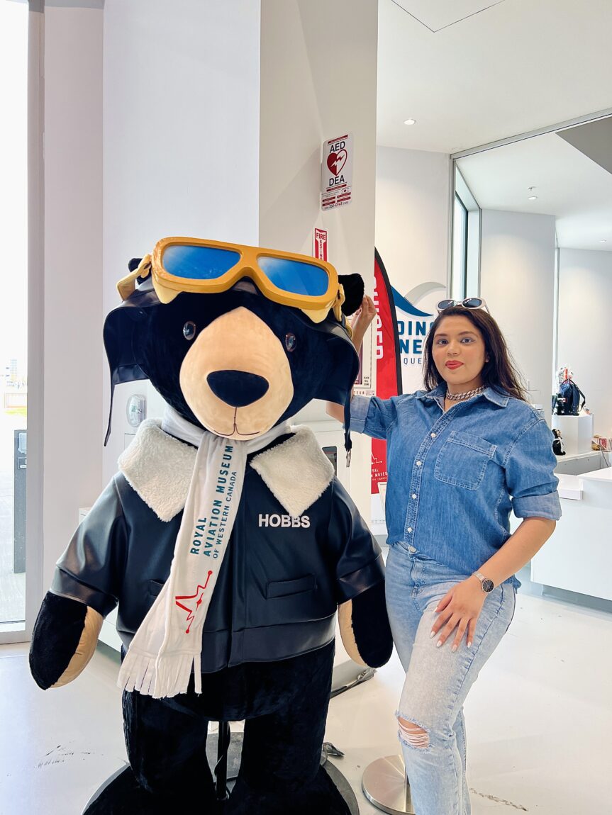 A female visitor at the Royal Aviation Museum stands next to the museum's mascot, Hobbs the Bear. The visitor is wearing a dark denim button up and light denim jeans. Hobbs is lifesize, wearing a leather aviator jacket, aviator goggles, and a white scarf bearing the RAMWC logo.
