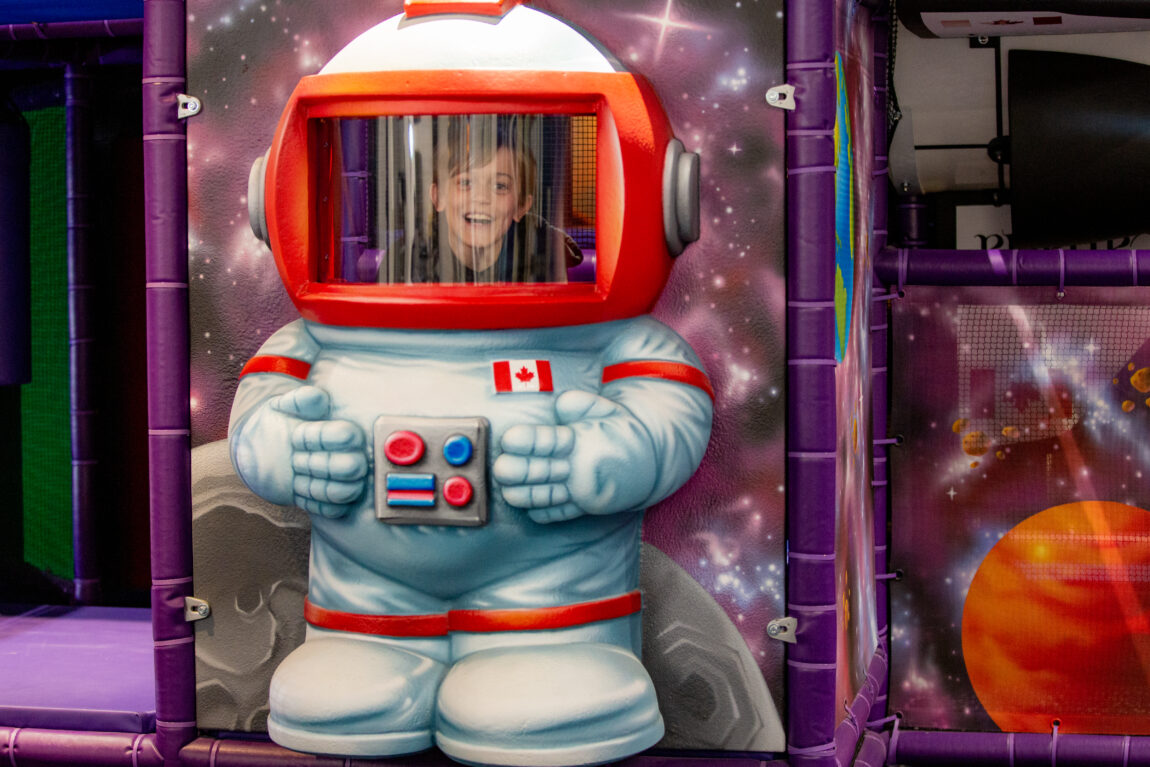 a young child peers out from inside an oversized astronaut's helmet in a space-themed play area