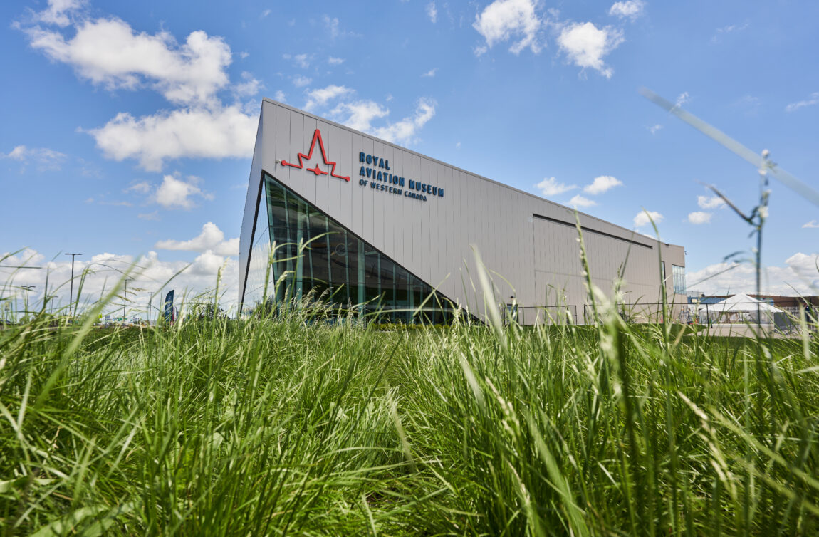 a brownish-grey building with a sign that reads 'Royal Aviation Museum of Western Canada' appears to rise out of the tall grass in front of it