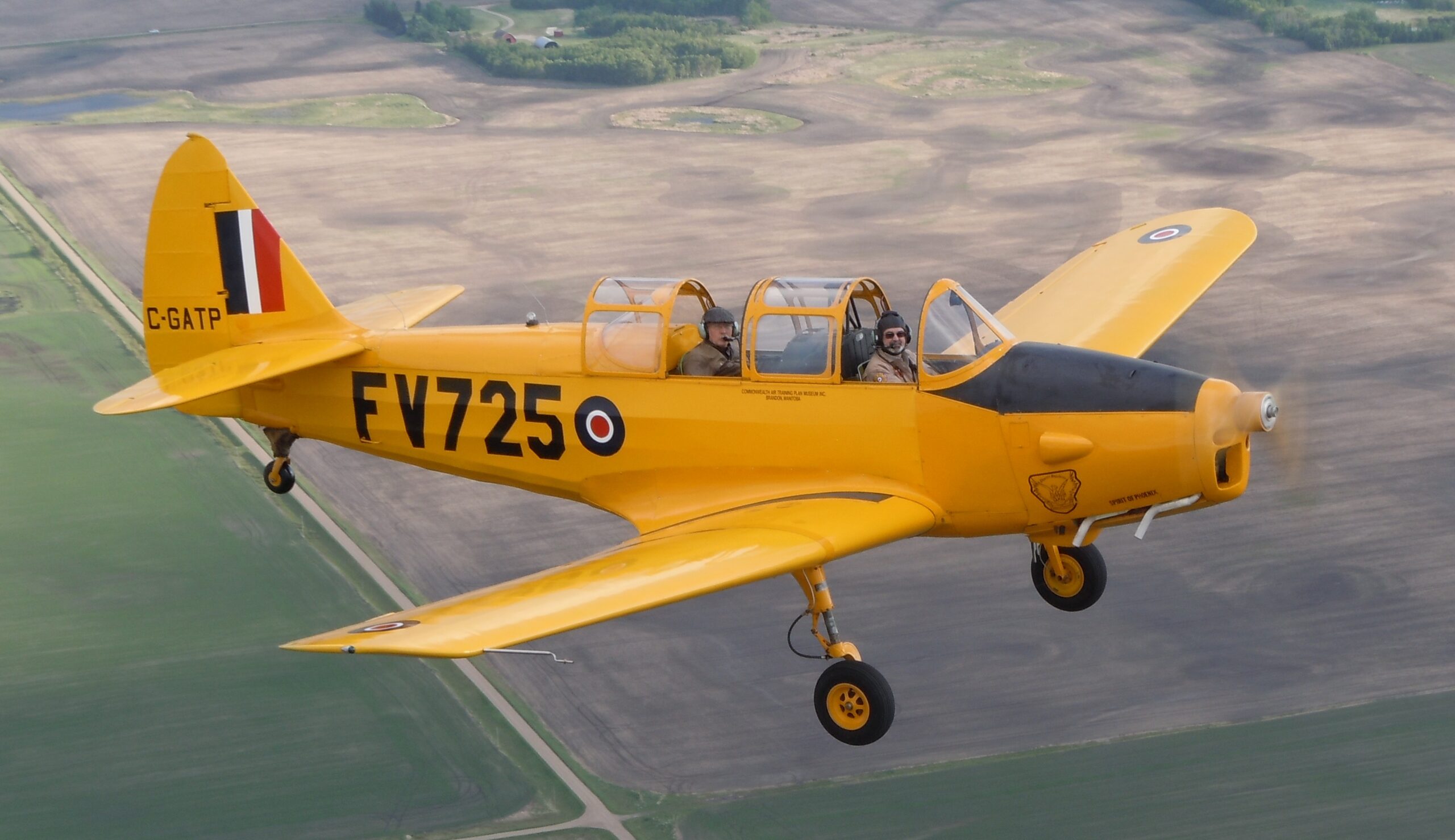 Fairchild Cornell PT26 aircraft flying above a patchwork of farmland. The aircraft is bright yellow, mostly fills the frame, and is seen from the right side. On its side, the call letters 'FV723' appear along with an RCAF roundel.