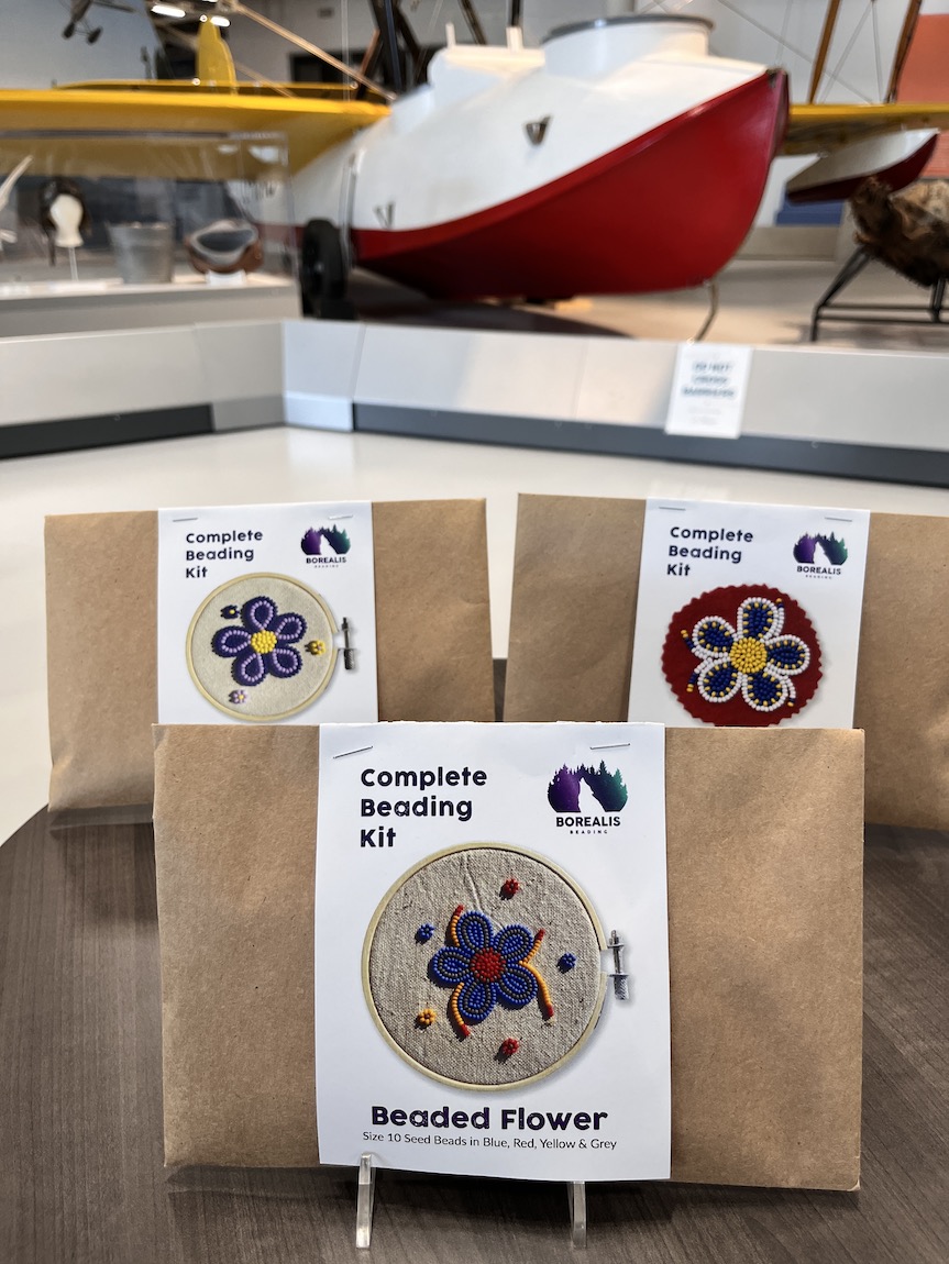Indigenous beading kits in brown craft paper on a small round table with an antique white, red, and yellow aircraft in the background