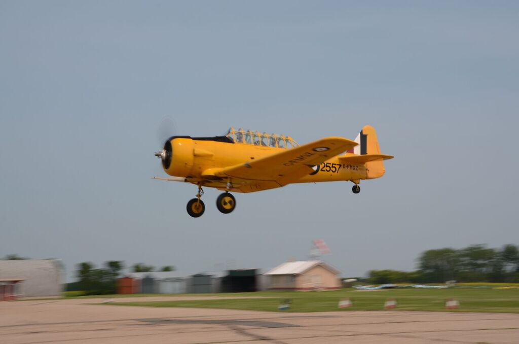 A North American Harvard Mark II aircraft flies low to the ground. The aircraft is yellow and in the background a few small buildings are visible.