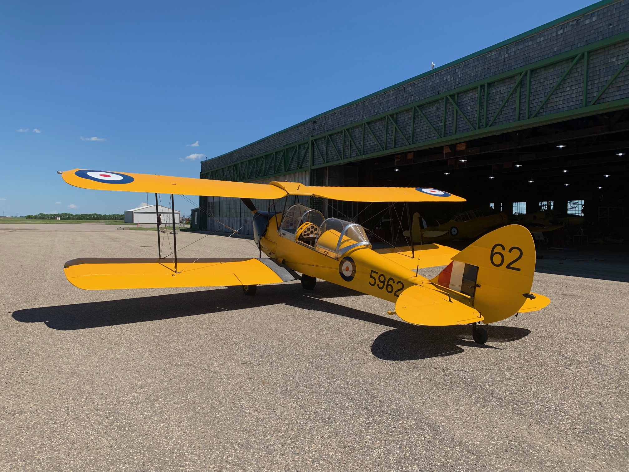 A Canadian DeHavilland Tiger Moth sits on the tarmac outside a hangar. The aircraft is bright yellow and decorated with RCAF roundels. On the tail, the number '62' appears.