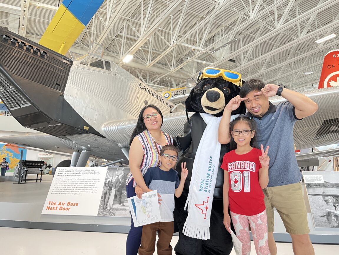 a young family poses with a museum mascot in front of a large cargo aircraft