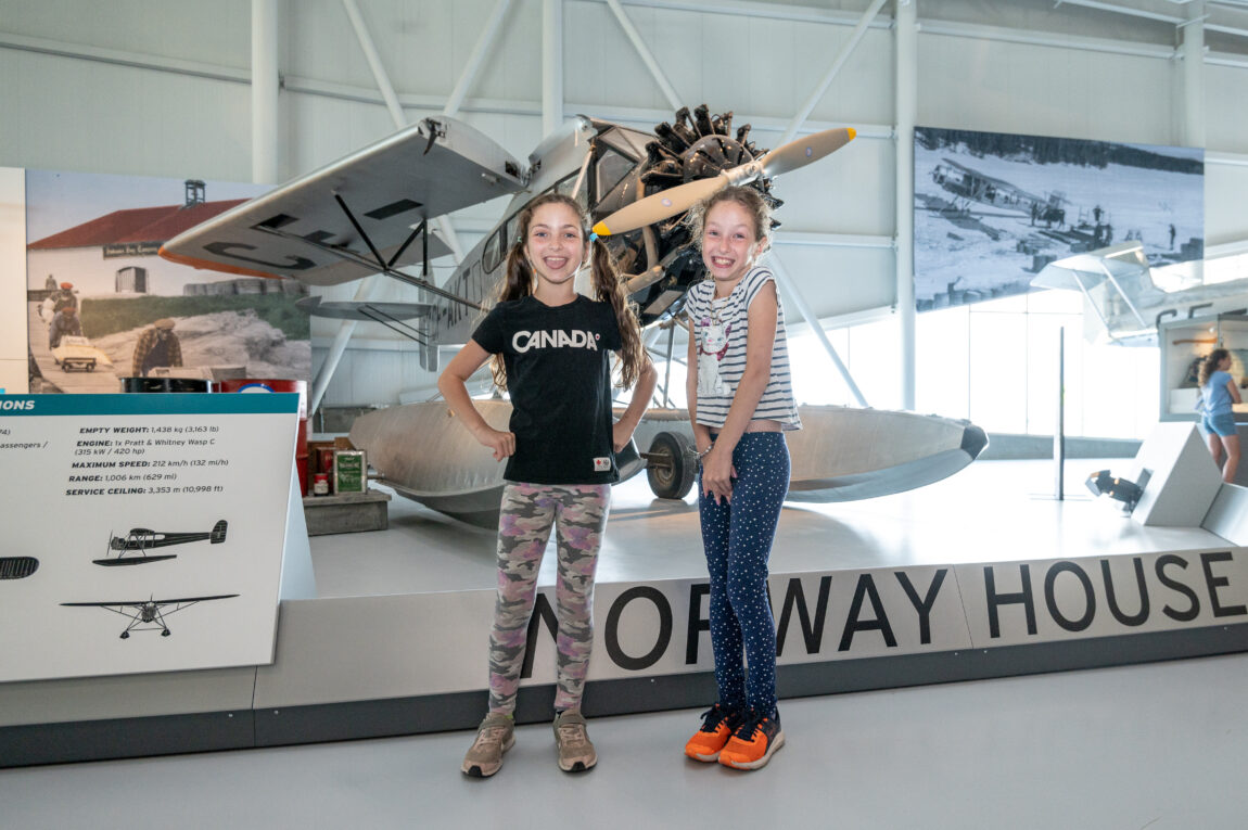 two young girls stand in front of a vintage aircraft in a museum