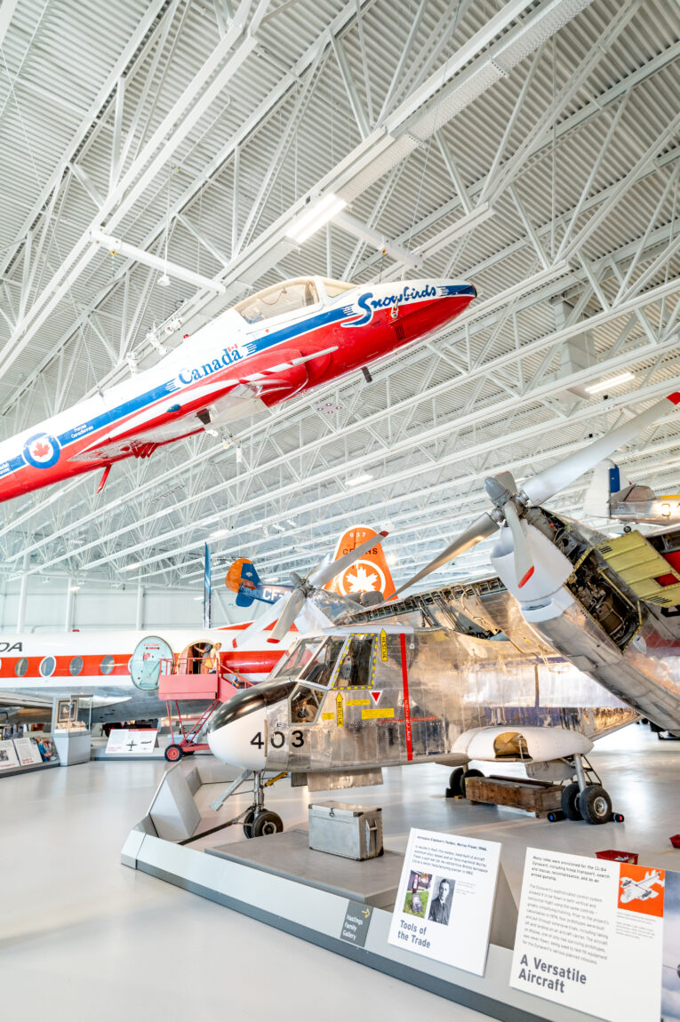 a Tutor Snowbird aircraft hangs suspended from a museum ceiling. Below it, other assorted aircraft fill the museum floor