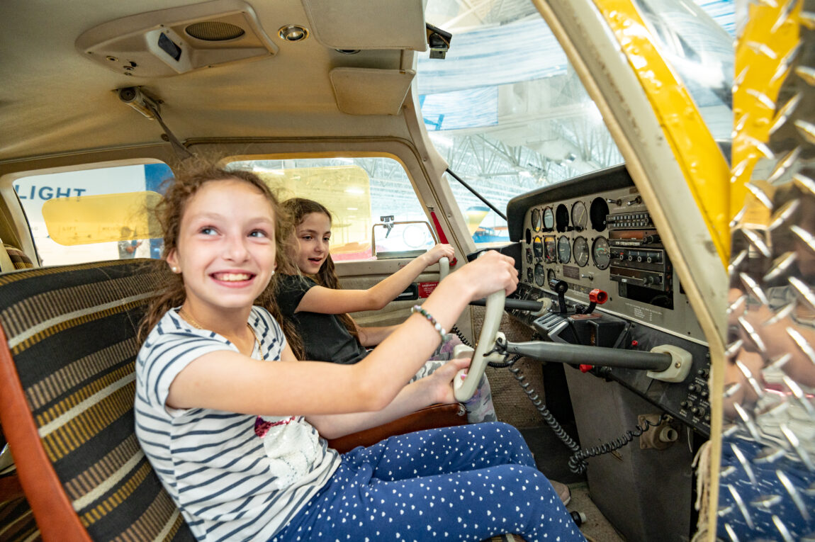 Two young girls sit at the controls in the cockpit of a small plane