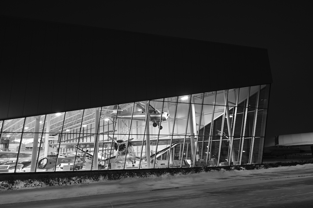 Exterior of a museum in black and white. Most of the exterior wall is windows and inside multiple aircraft are visible