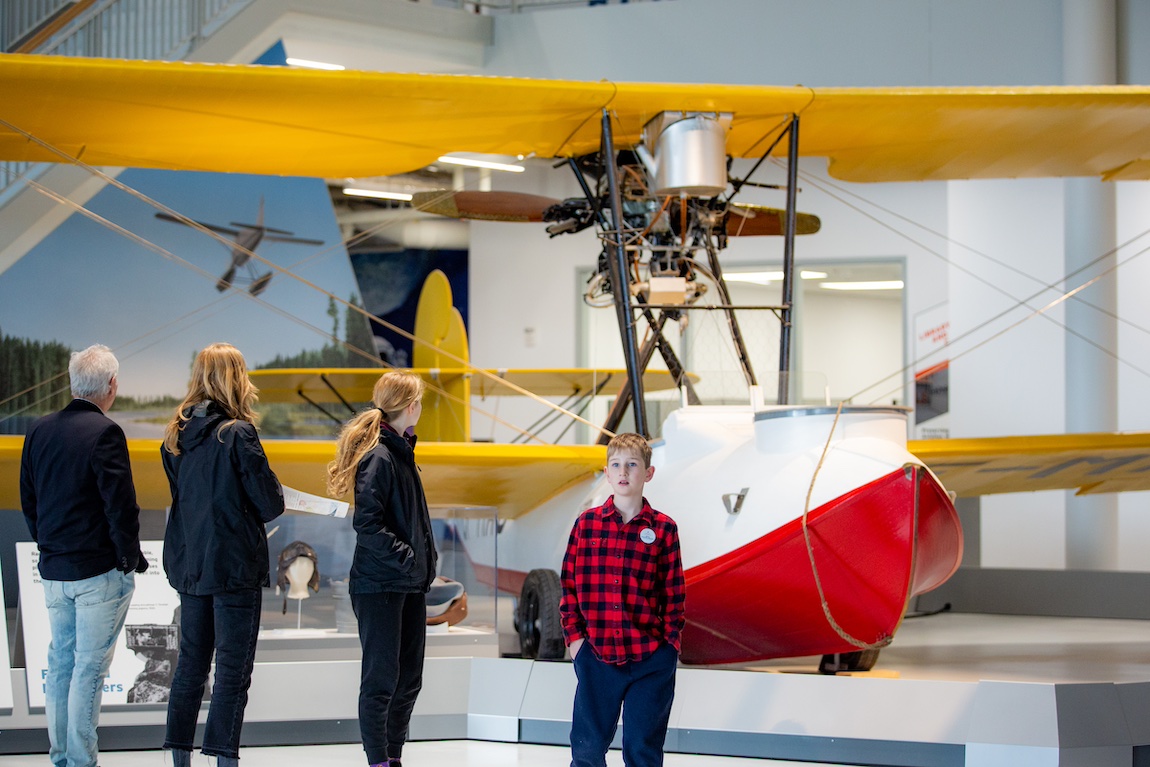A family stands in front of a vintage red and white Vickers Vedette flying boat with red wings. Their backs are to the camera with the exception of a young boy who is looking at the camera.