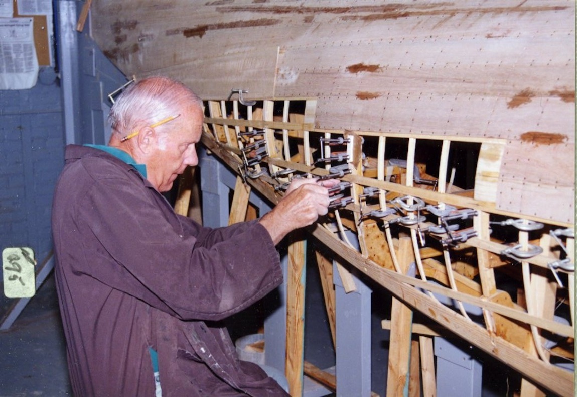An elderly gentleman affixes clamps to the bottom of a boat hull being built