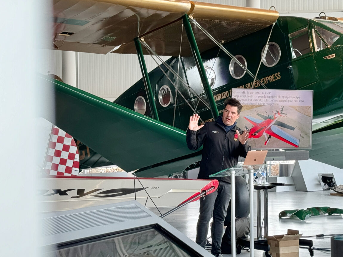 A man stands at a podium in front of a vintage green aircraft giving a presentation