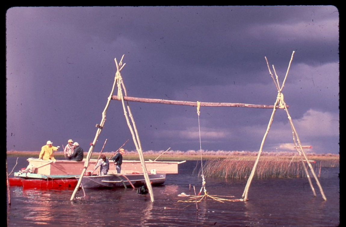 Two A-frame structures are in a lake with a long log connecting them at the top. From the log hands a rope that appears to be pulling part of a boat wreck out of the water.