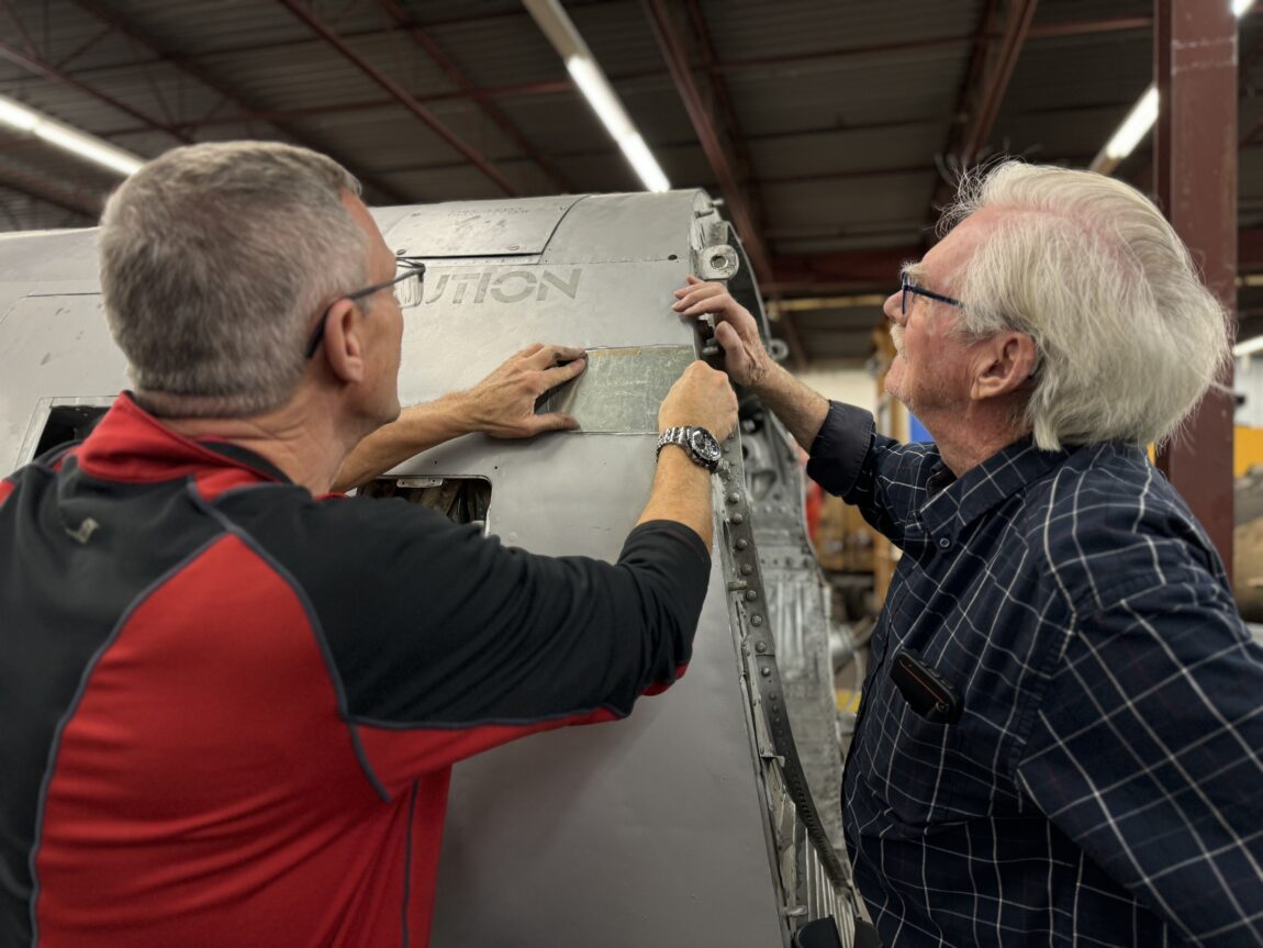 Two men affix a patch to the fuselage of an aircraft under restoration