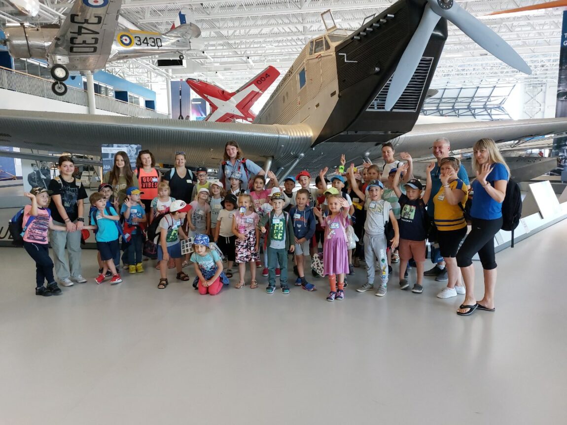 Summer Fun children's programming participants post in front of a large, antique cargo plane inside a hangar-style building. 
