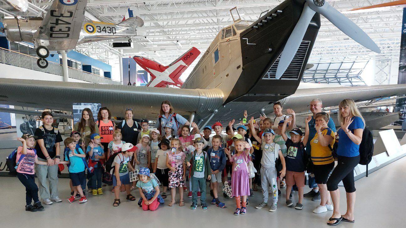 A group of excited children participating in summer programming pose in front of a vintage cargo plane that's parked inside an aviation museum.