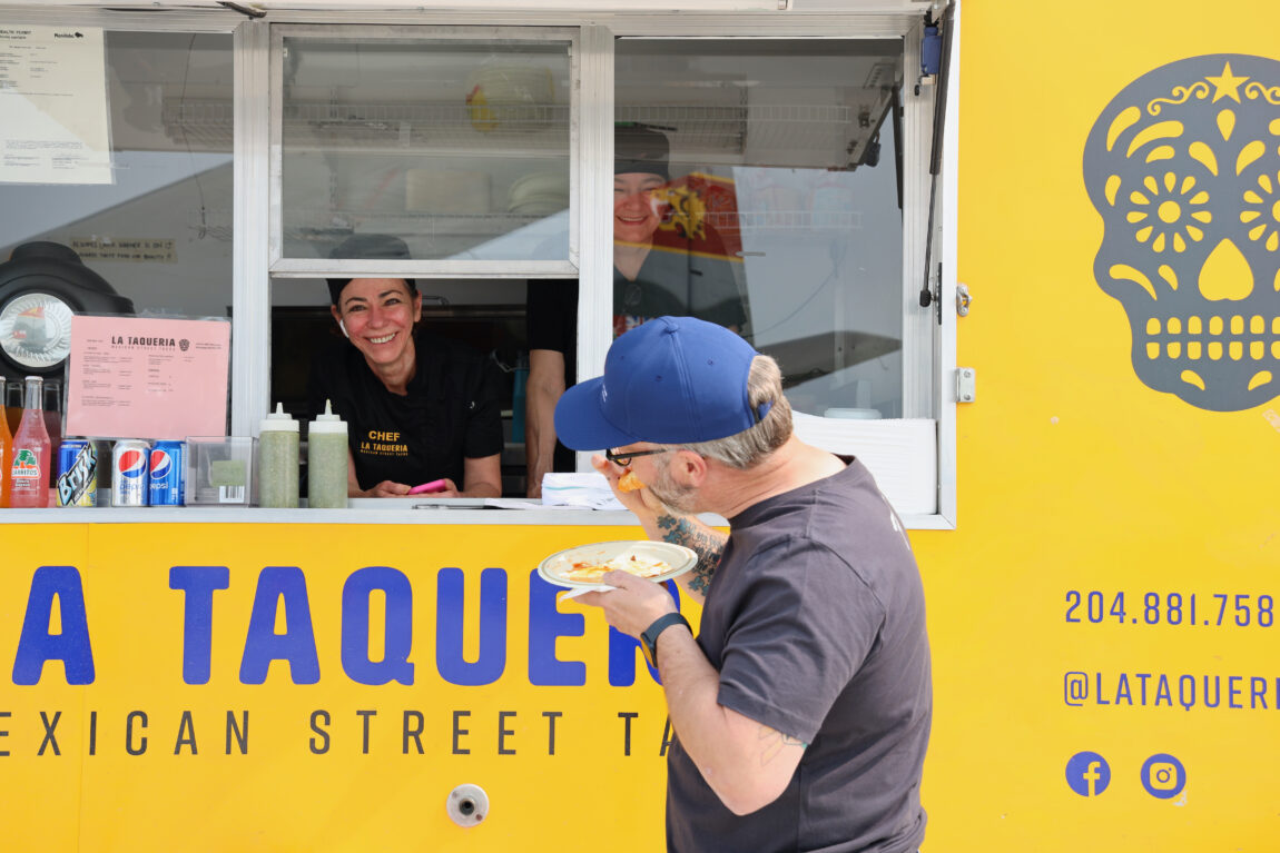 A man stands in front of a yellow taco truck, taking a bite of his food. Inside the truck, the chef smiles out at the camera.