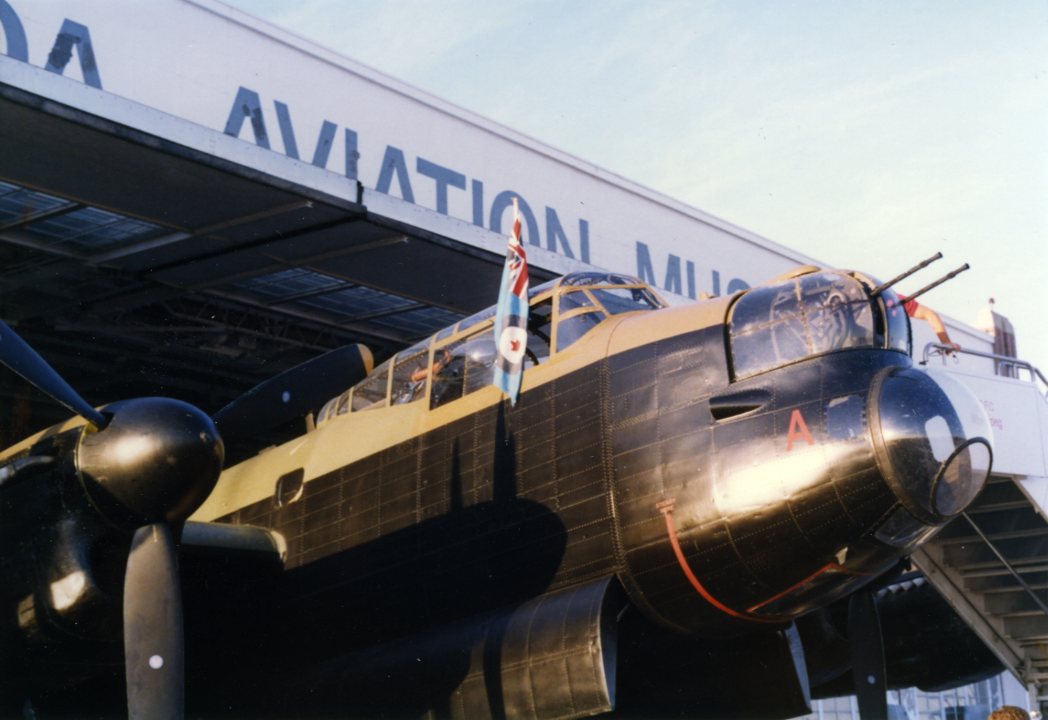 Avro Lancaster parked in front of a hanger with the name, 'Western Canada Aviation Museum' partly visible. The nose and the aircraft is visible in front of the hangar door.