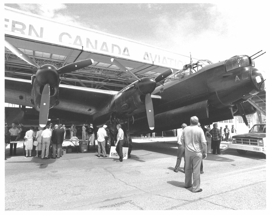Avro Lancaster parked in front of a hanger with the name, 'Western Canada Aviation Museum' partly visible. The nose and right wing of the aircraft are seen in front of the hangar door.